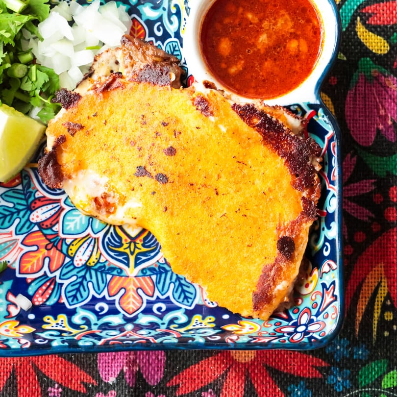 Close-up of a golden-brown quesabirria taco with melted cheese, cilantro, onions, lime, and consommé.
