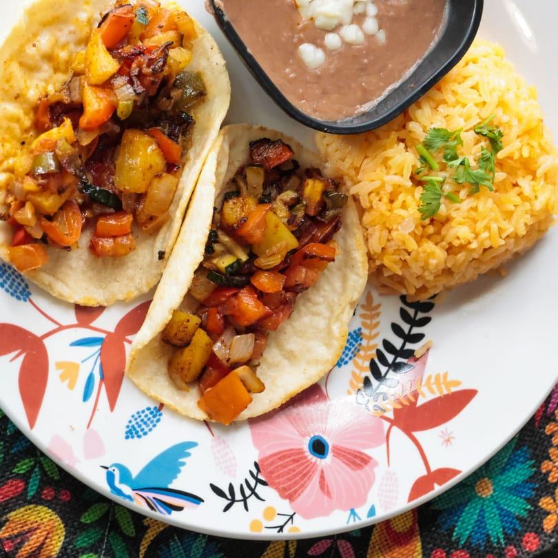 Two vegetable tacos with rice and refried beans on a colorful floral plate.